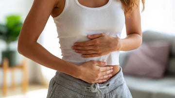 Mujer con camiseta blanca sin mangas y pantalones de chándal grises sujetándose el abdomen con ambas manos debido a calambres o dolor de estómago